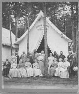 SA0159 - A group of men and women in front of tent in a grove, showing a campground setting.